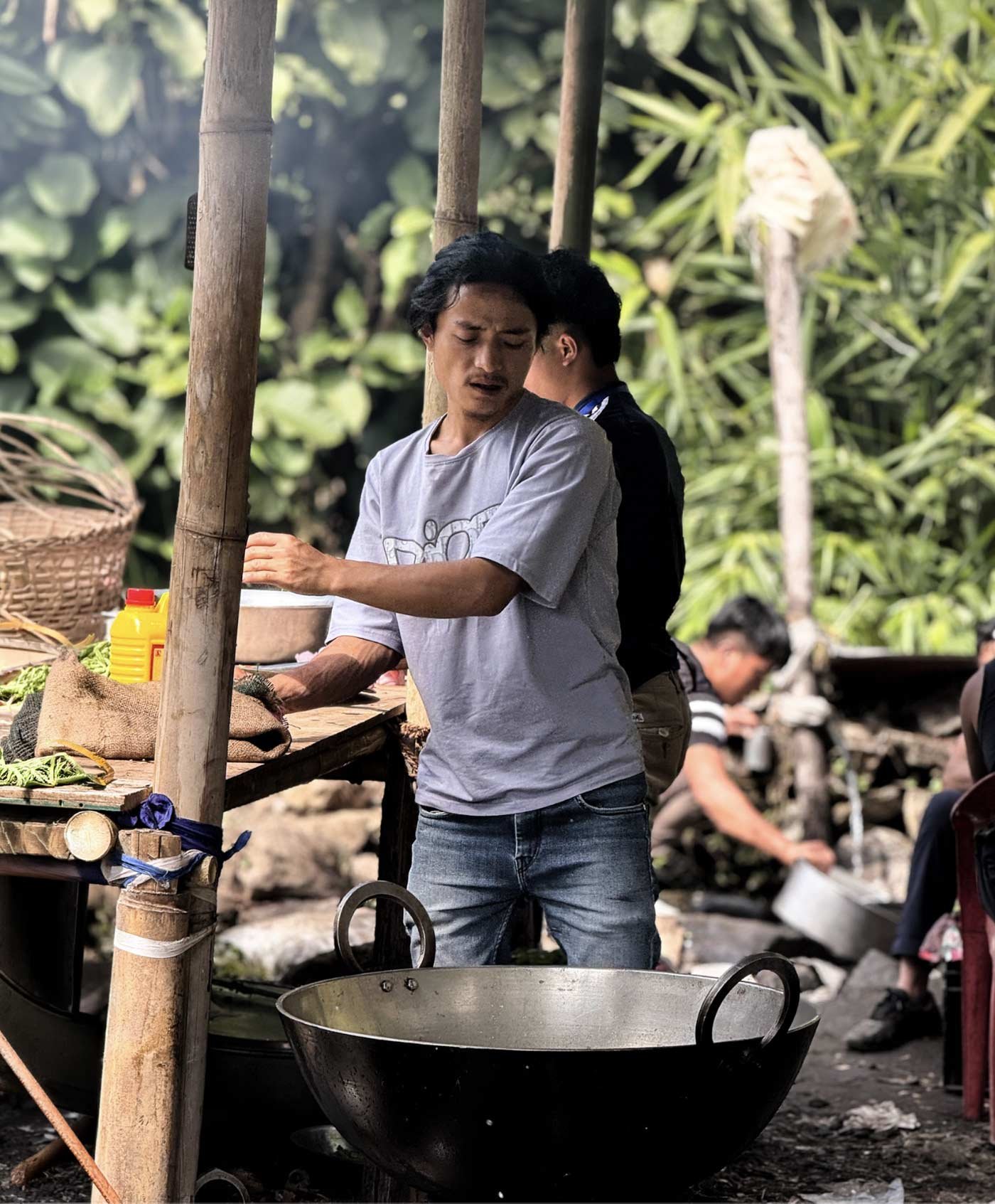 A man in a gray t-shirt and jeans cooking outdoors at a rustic food stall with a large black wok, with lush greenery in the background and other people around.