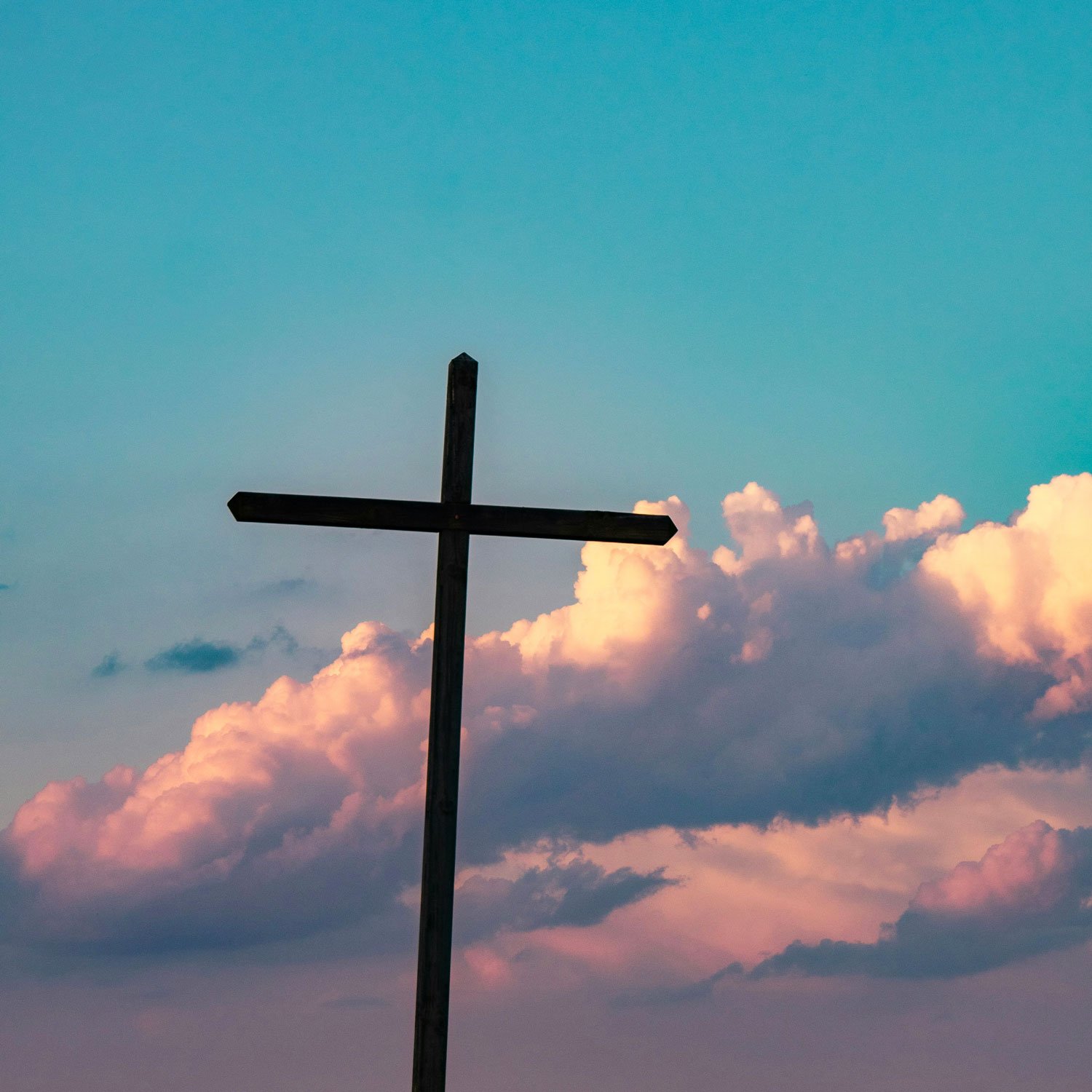 A wooden cross silhouette against a colorful sky with pink and orange clouds at sunset or sunrise.