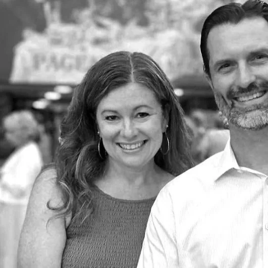 A smiling woman with wavy hair wearing a sleeveless top standing next to a smiling man with dark hair and a beard wearing a collared shirt, in a social setting with blurred background.