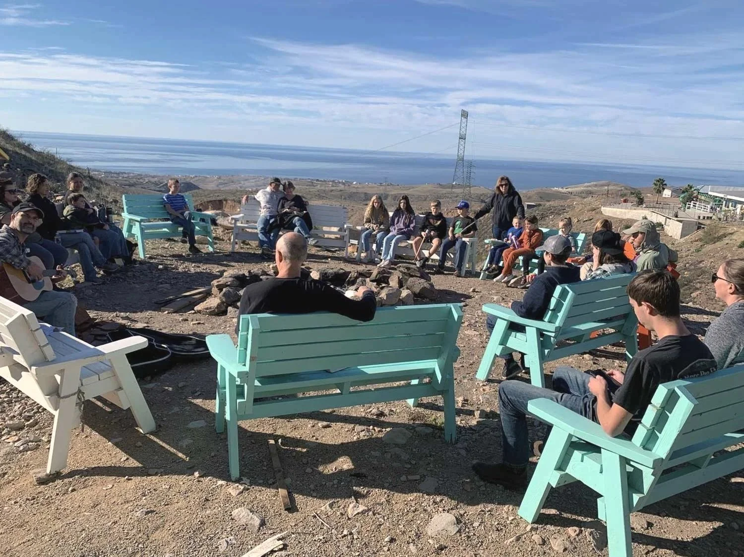 Group of people gathered around a campfire on a hillside with ocean view, some sitting on colorful benches, some playing guitars, with clear sky and distant landscape in the background.