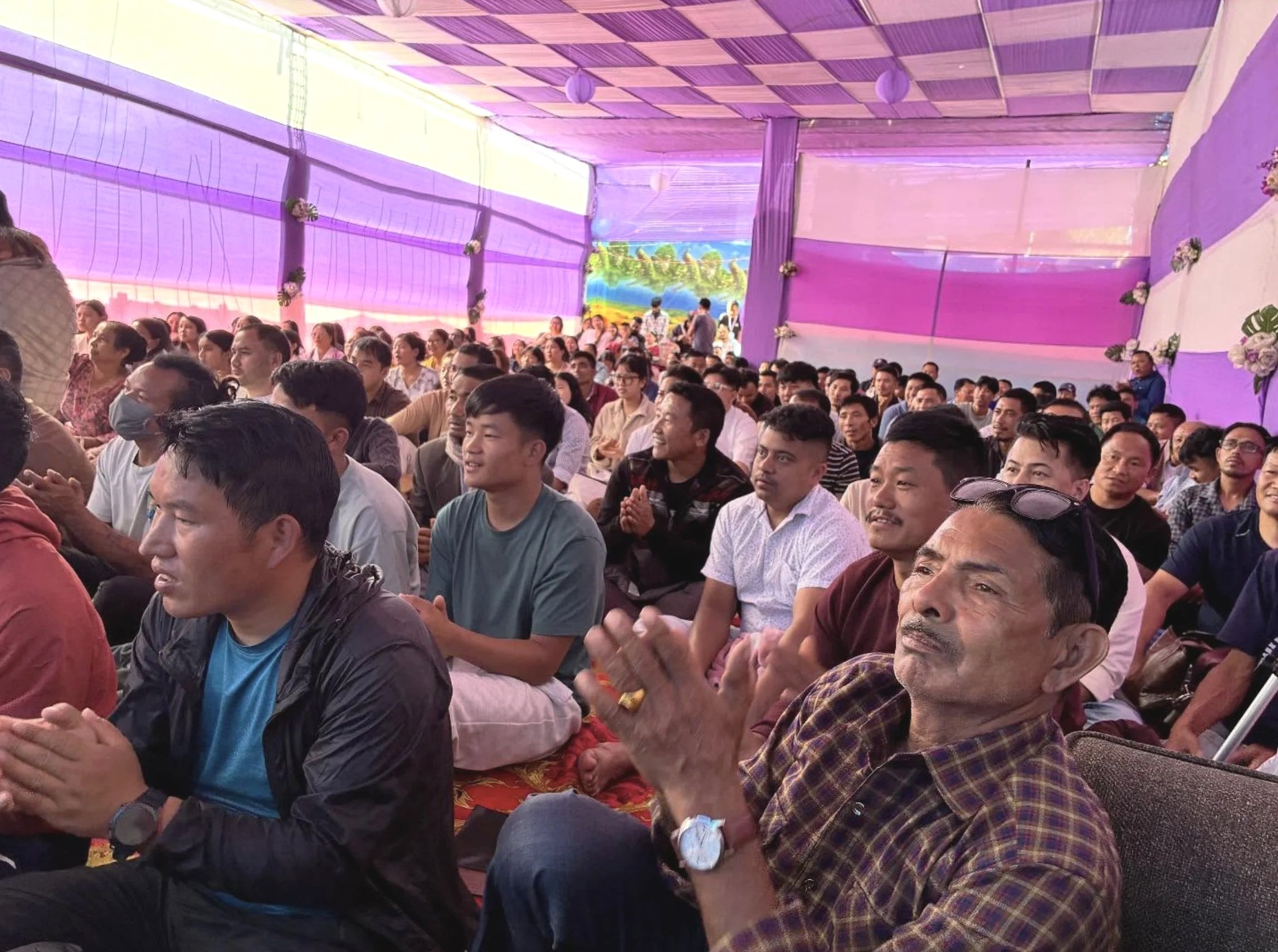 A crowd of people sitting together inside a decorated tent with purple drapes and floral arrangements, attending an event.