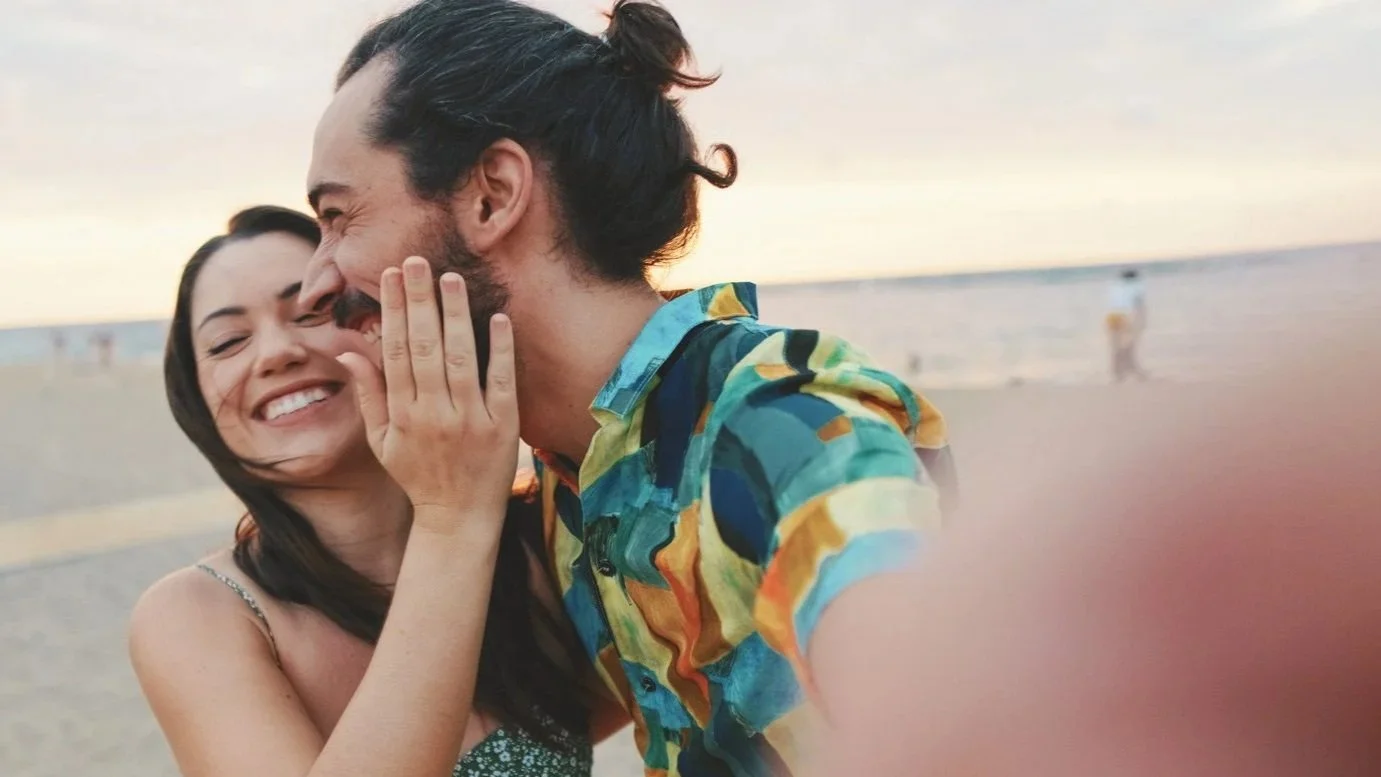 A young couple taking a selfie at the beach during sunset, both smiling and laughing tightly together.