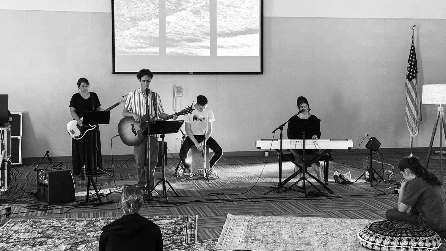 Black and white photo of a musical performance in a classroom or lecture hall. Four young women and one young man are on stage, with three playing instruments and one singing into a microphone. The instruments include a guitar, a bass, a cajón, and a keyboard. Two people in the audience are sitting on the floor, listening attentively.
