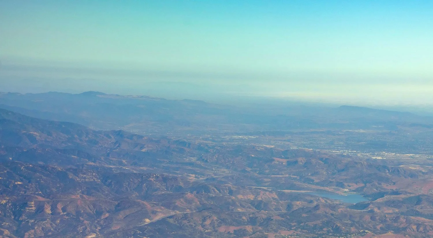 Aerial view of a mountain range with a body of water and distant plains under a clear sky.