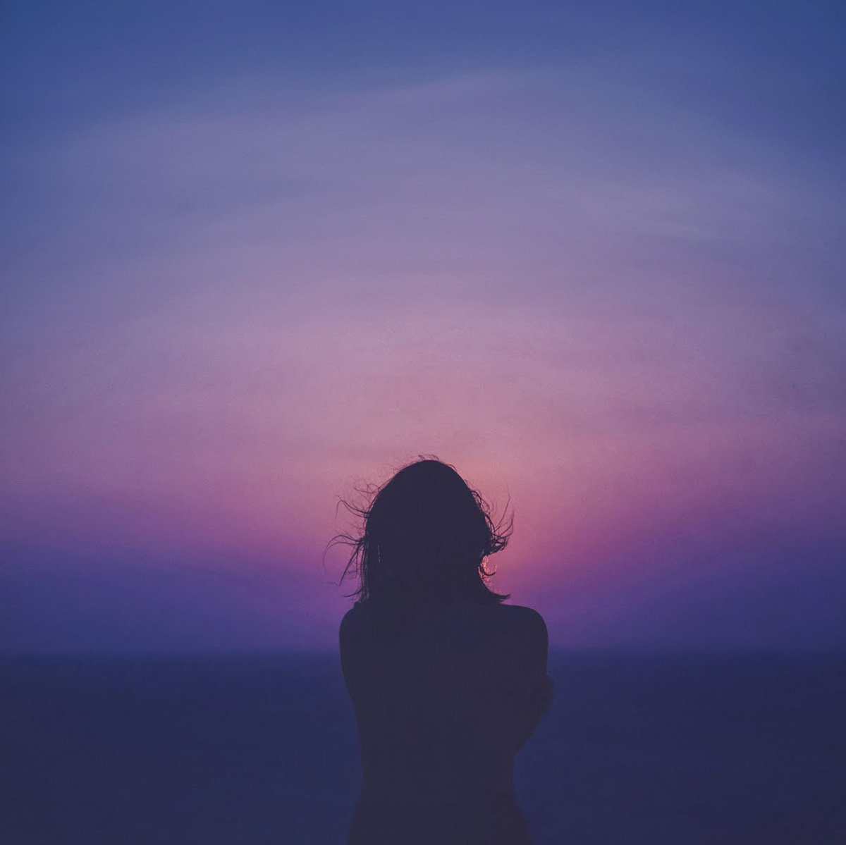 Silhouette of a woman with windblown hair standing against a colorful sunset sky over the ocean.