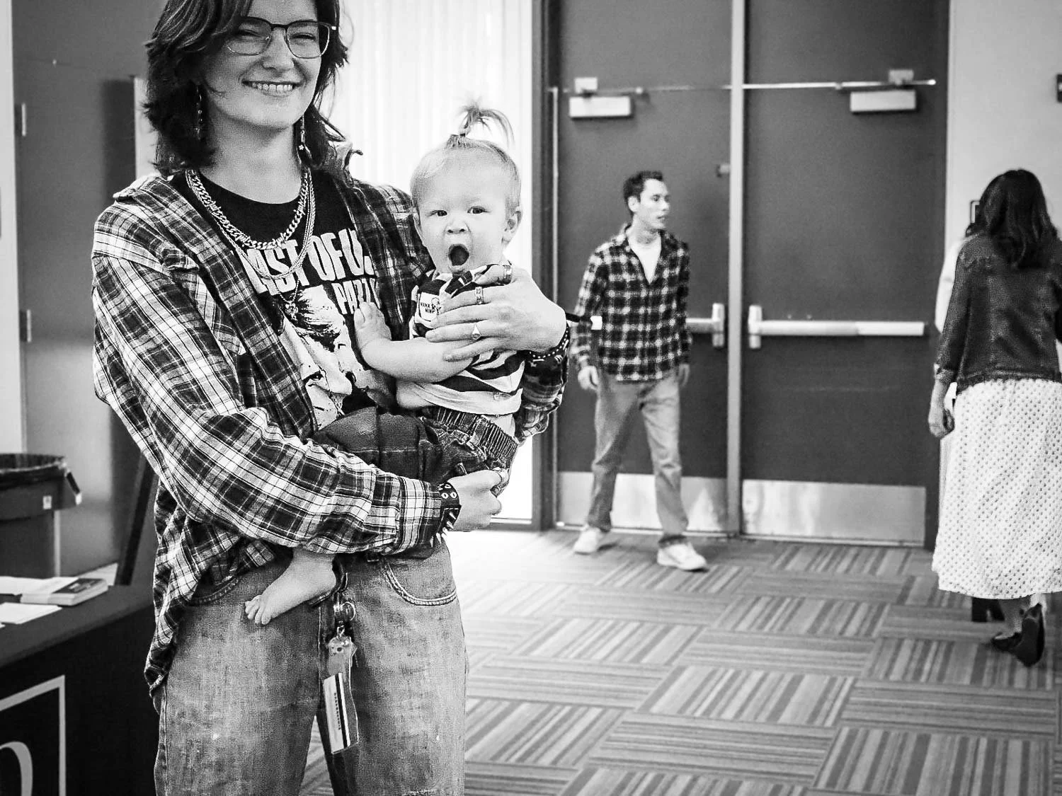 A woman with glasses and layered necklaces is smiling and holding a young child who appears to be yawning. The woman is wearing a plaid shirt over a graphic T-shirt and jeans, with keys hanging from her belt. In the background, two people are near a set of double doors in what looks like an indoor public space.