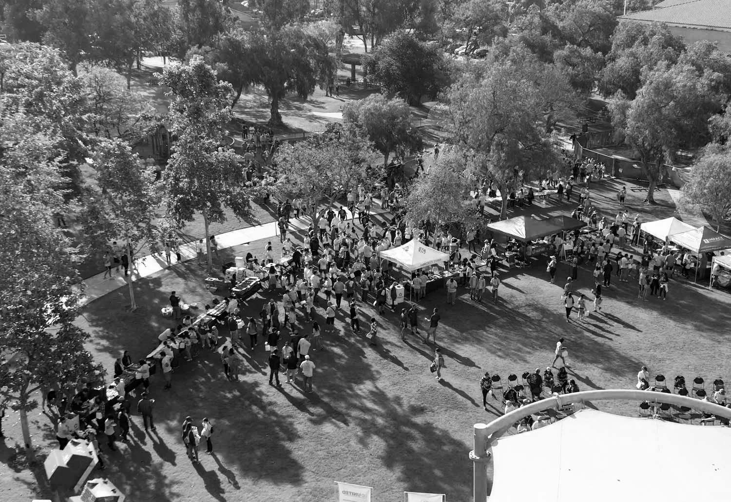 A crowd of people gathered outdoors at a fair or market with trees and tents.