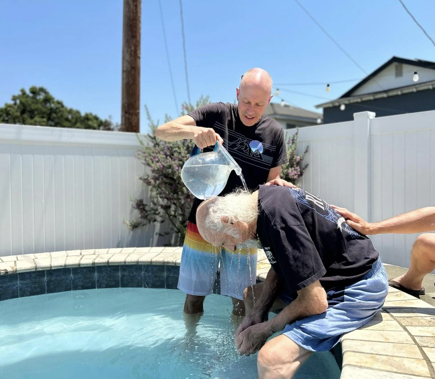 A man pouring water on another man leaning into a pool, with a third person gently holding his shoulder. They are outdoors under clear blue skies, with a white fence and some bushes in the background.