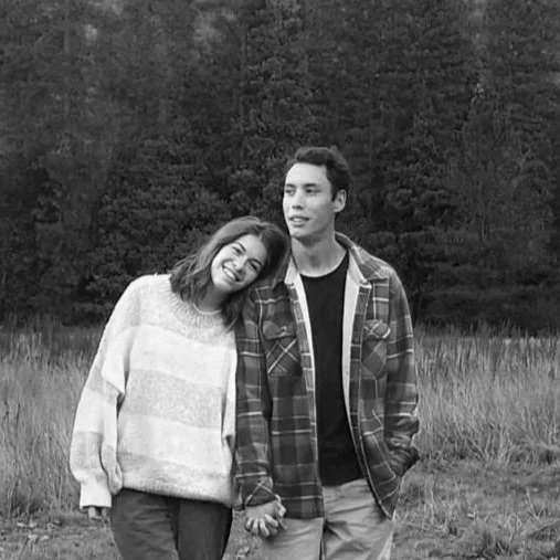 A smiling woman leaning her head on a smiling man holding hands outdoors, with trees and mountains in the background.