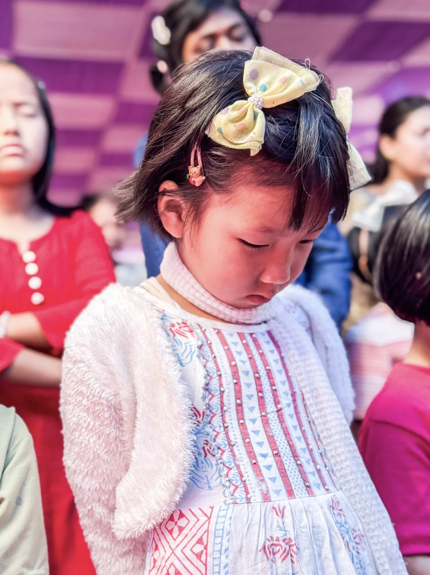 A young girl with short dark hair wearing a large yellow bow with colorful polka dots and a pink bear hair clip, dressed in a white textured sweater and a dress with blue, pink, and white patterns, is looking down with others blurred in the background.