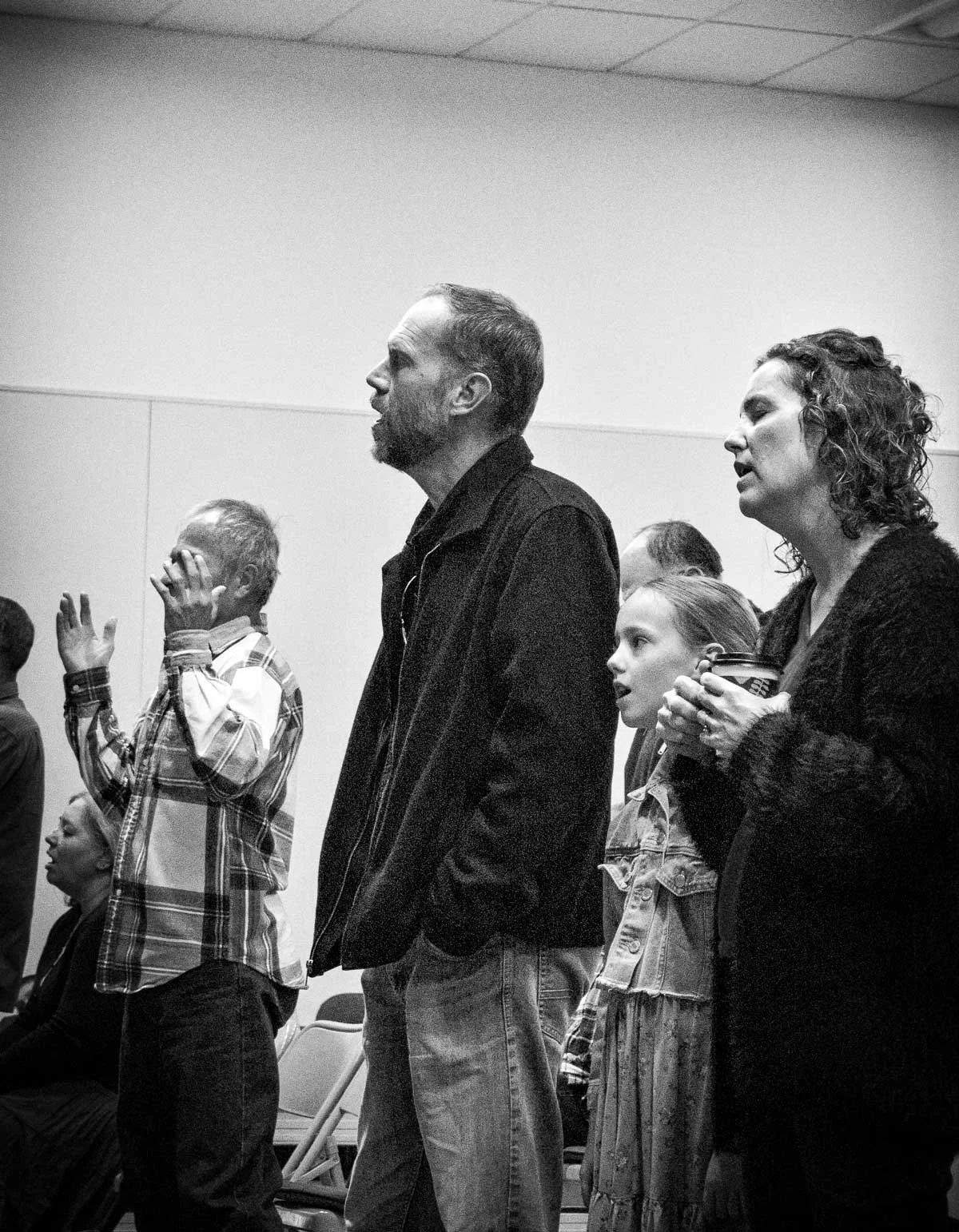 Group of people standing indoors, some with eyes closed or hands raised, appearing to be praying or participating in a moment of reflection.