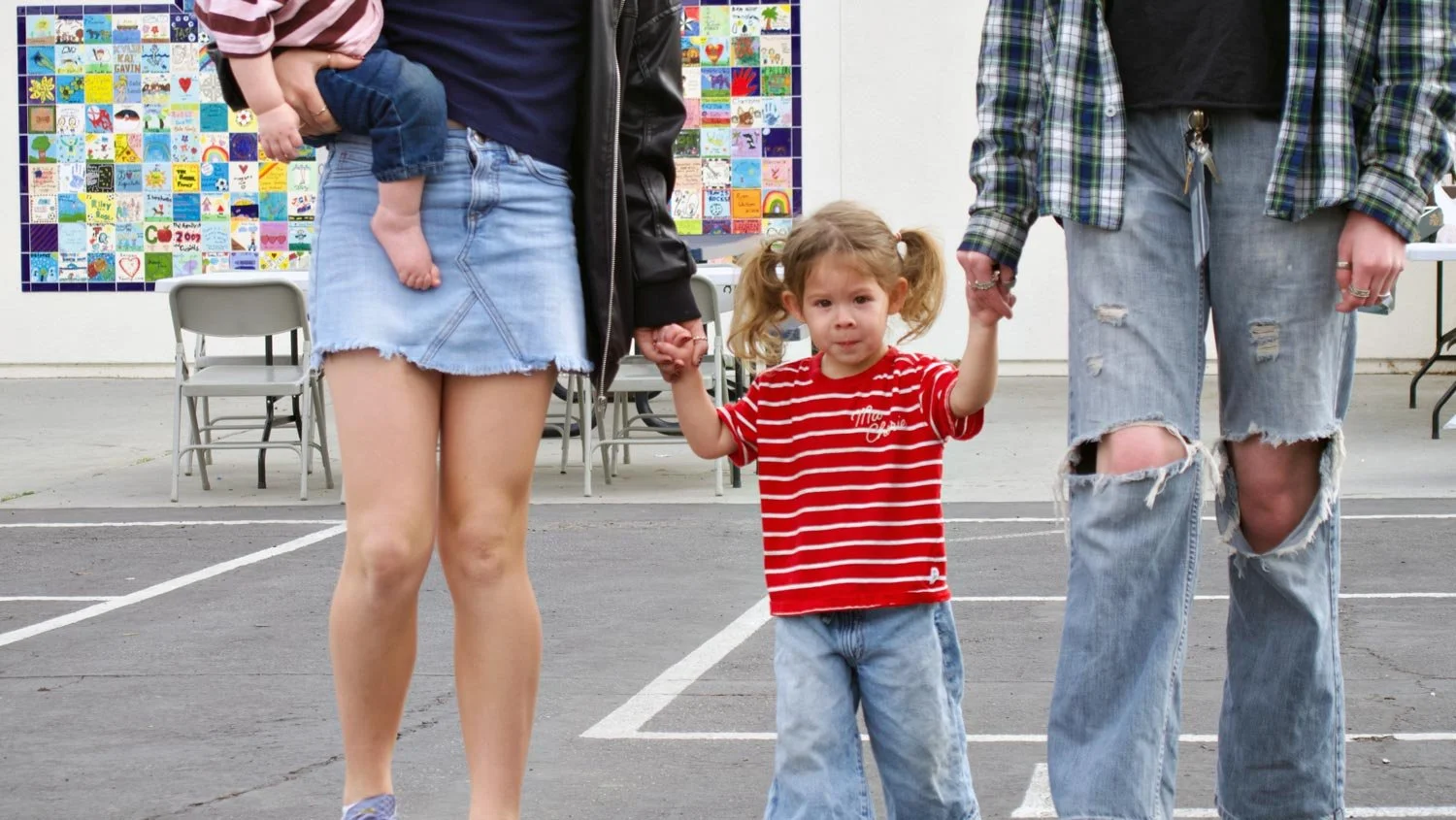 A young girl with pigtails and a red striped shirt holding hands with two adults, one on each side, in a parking lot with chairs and a colorful mural in the background.