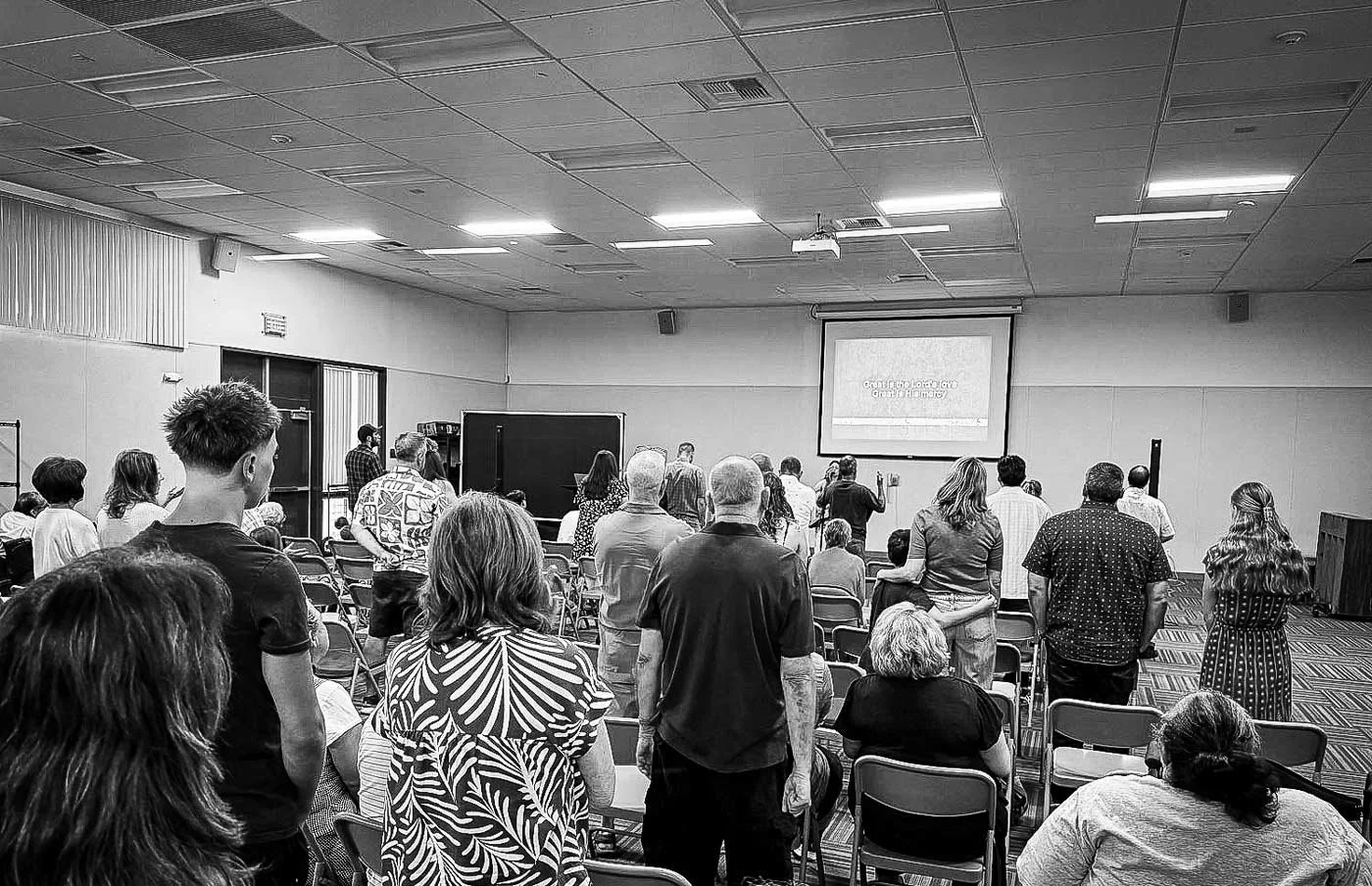 People standing and sitting in a large conference room facing a presentation screen.