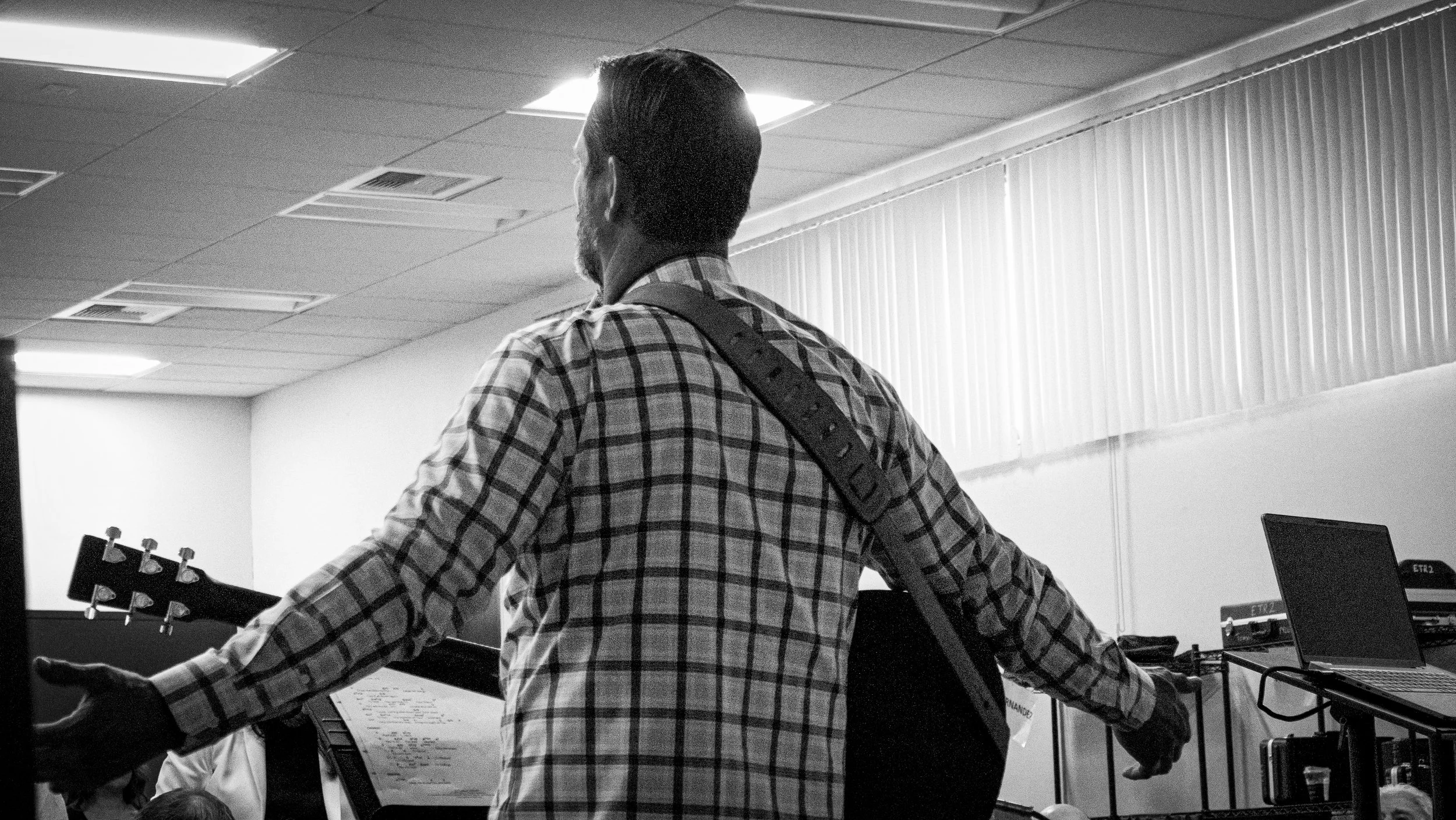 A person with short hair and a beard wearing a checkered shirt, standing in a room with musical equipment and a music stand, facing away from the camera.