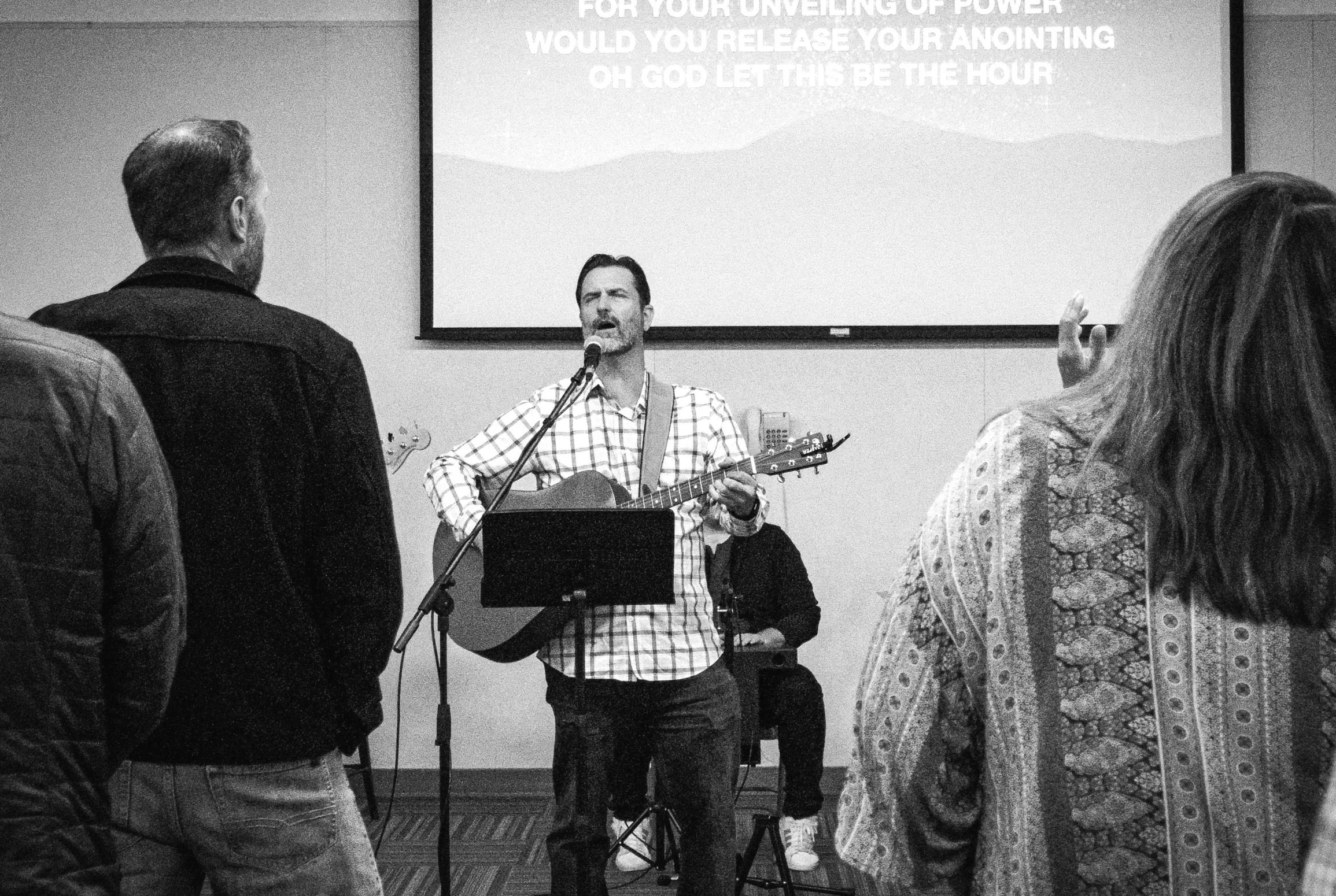 A man playing a guitar and singing into a microphone during a religious gathering, with an audience listening. A screen behind him displays partial text related to God's power and anointing.