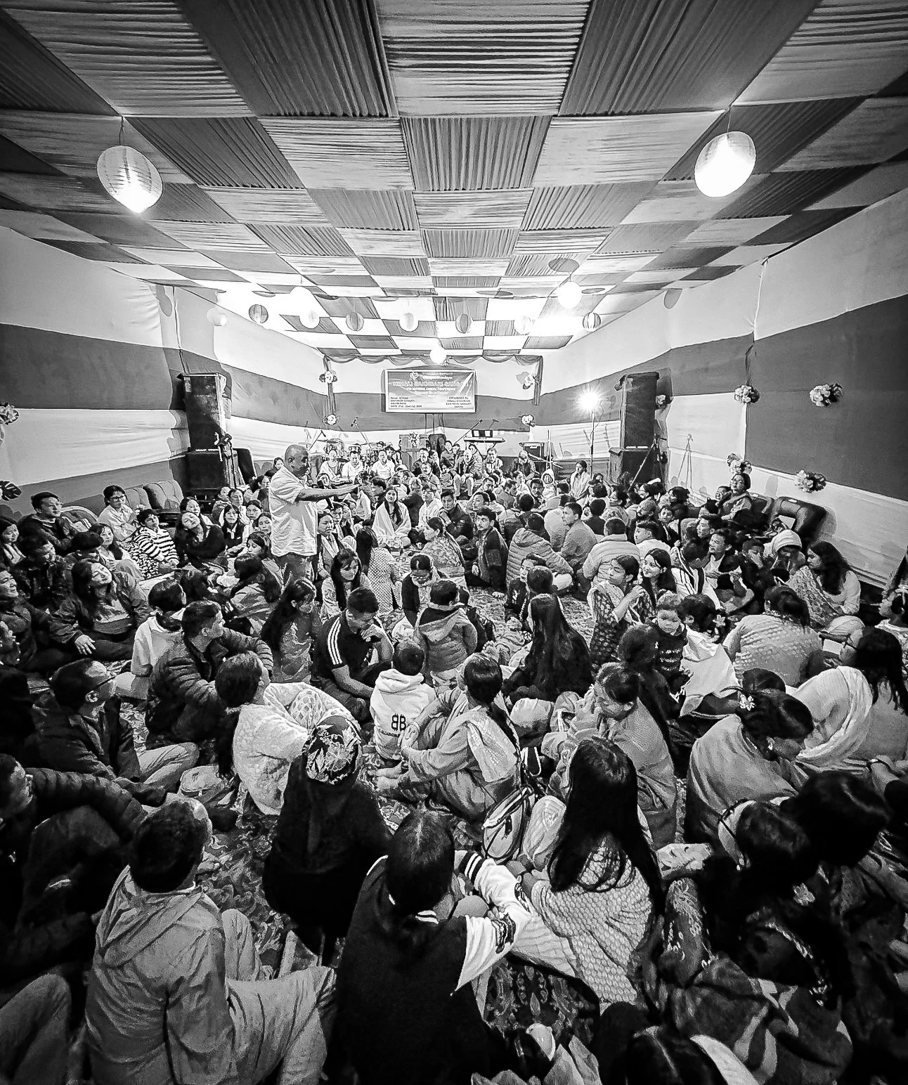 A large indoor gathering of people sitting on the floor, listening to a speaker standing in front of a stage, with a decorated wall and speakers on either side.