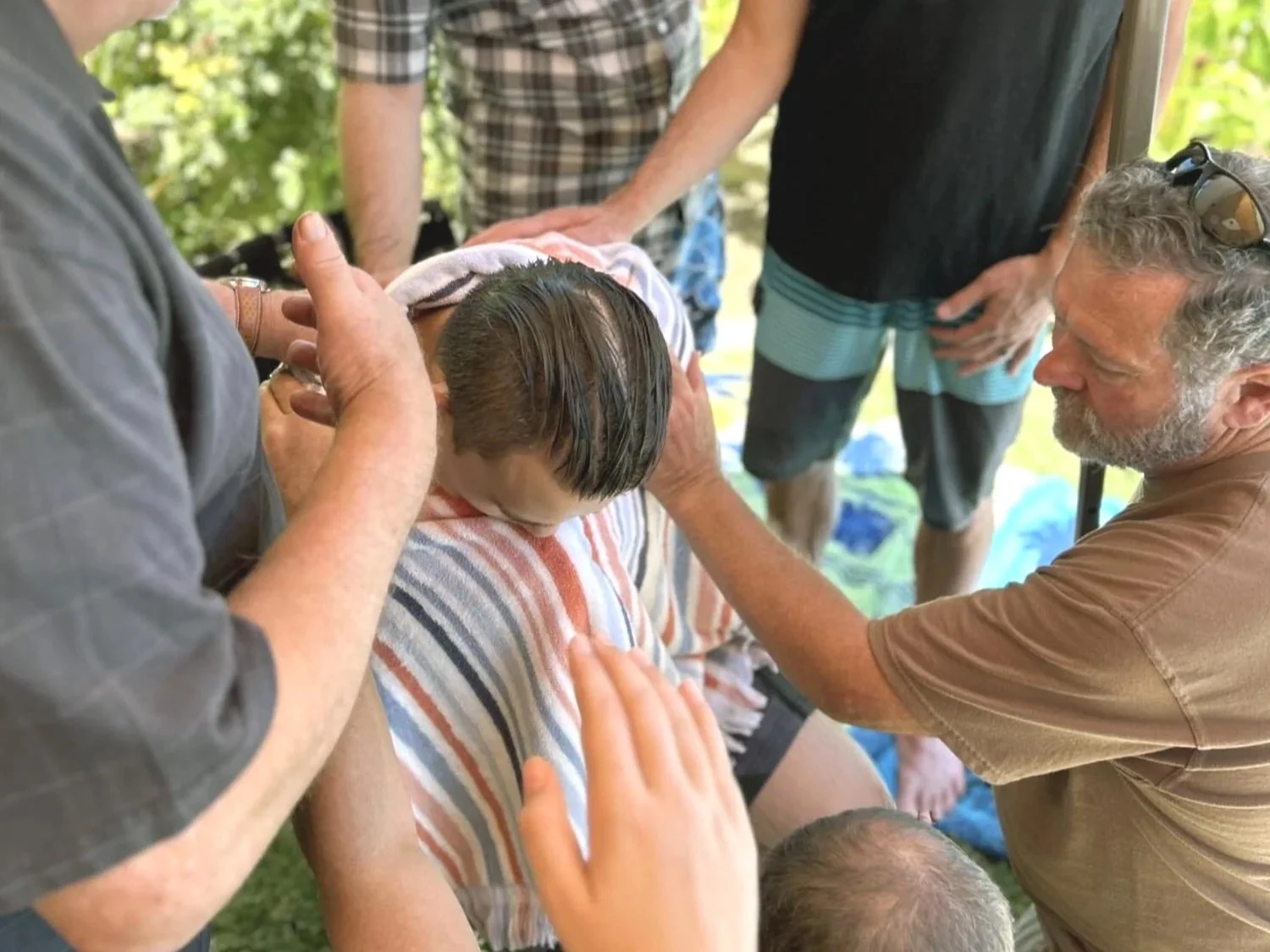 Group of people surrounding and praying over a young boy outdoors during a religious ceremony.