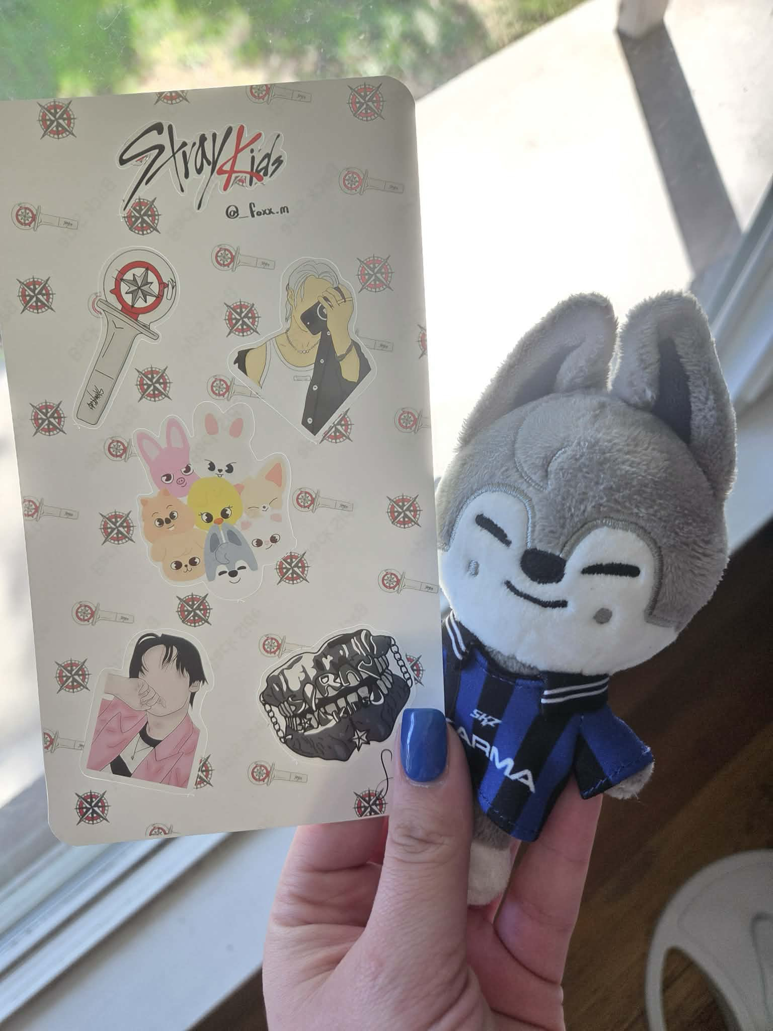A hand holding a piece of paper with various stickers next to a plush toy of a wolf wearing a sports jersey. The paper has stickers of a sword, a person taking a photo, group of animal characters, a person with a hand covering their face, a chain with teeth, and a compass symbol. The background shows a window with sunlight.