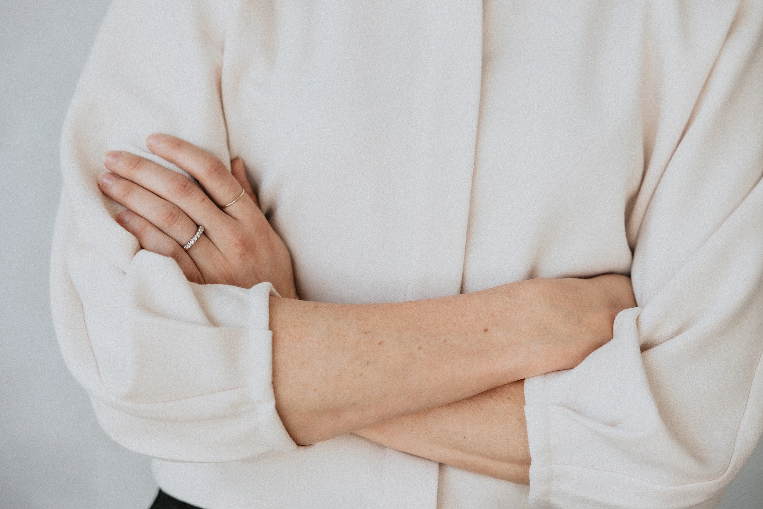 Close-up van een vrouw met gekruiste armen, gedragen in een crèmekleurige blouse, met sieraden aan haar vingers.