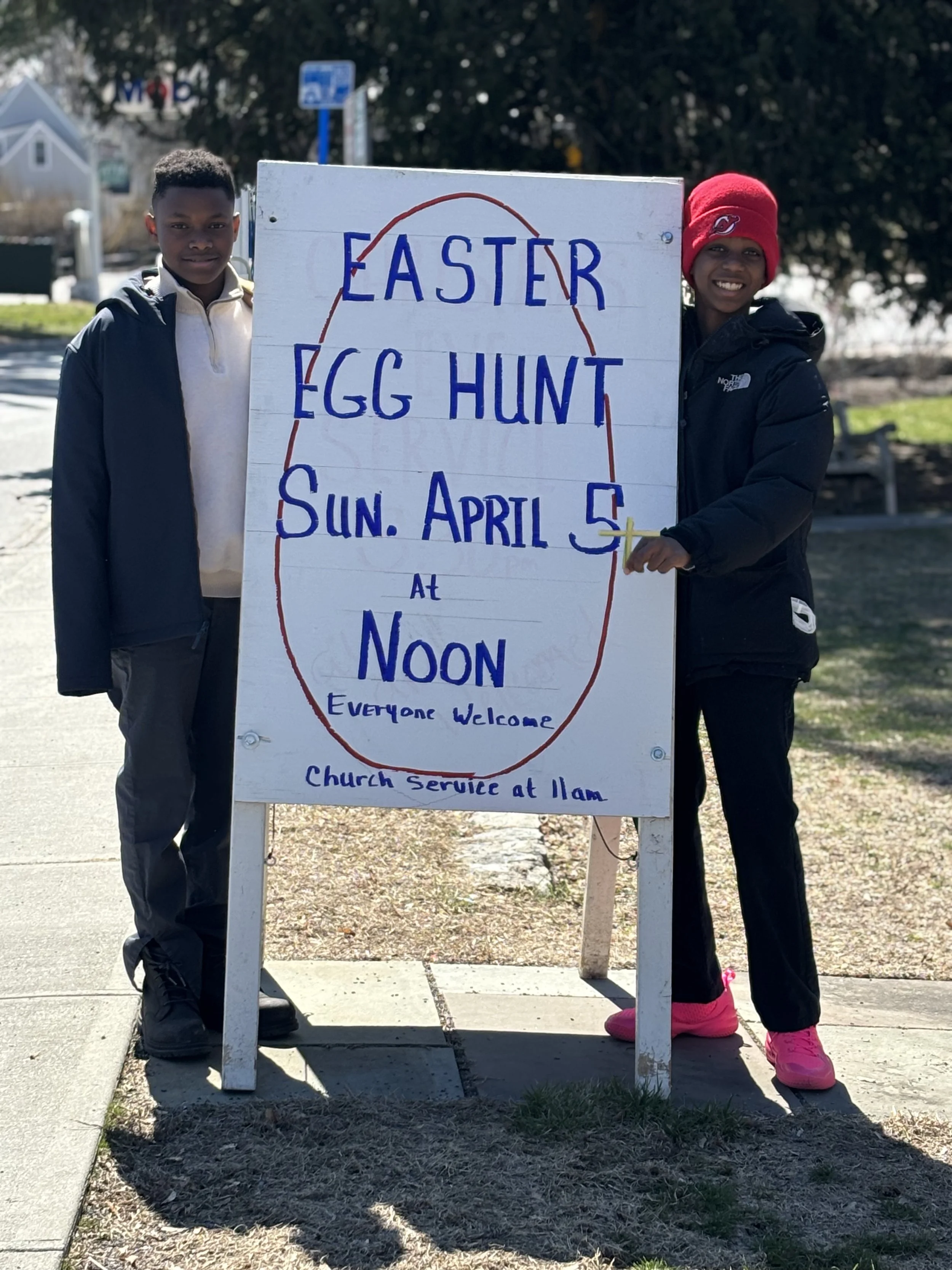 Two children holding a large poster sign that reads 'EASTER EGG HUNT Sun. April 5 at Noon Everyone Welcome Church Service at Ilam' on a sidewalk. The boy on the left wears a black jacket and beige shirt, and the girl on the right wears a red beanie, black jacket, black pants, and pink shoes.