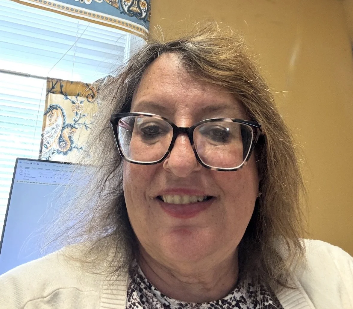 Close-up of a woman with glasses smiling indoors, with a computer monitor and window with curtains in the background.