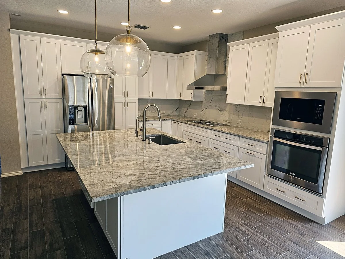 Modern kitchen with white cabinets, marble countertops, stainless steel appliances including a refrigerator, oven, microwave, and range hood. A kitchen island with a sink and pendant lights above. Dark wood flooring.
