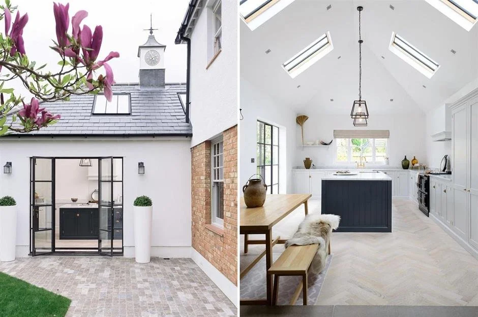 Side-by-side images of a modern home exterior and interior. The exterior shows a white house with a slate roof, large black-framed glass doors, and purple flowering branches in the foreground. The interior image displays a bright, airy kitchen with white cabinetry, a navy-blue island, a wooden dining table with a bench, and skylights in the vaulted ceiling.