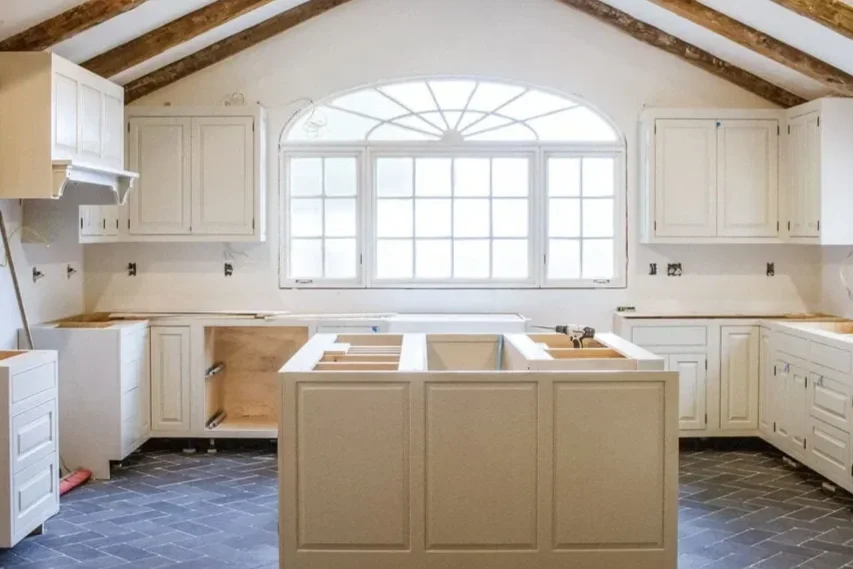 Kitchen with white cabinets, an island, large central window, and exposed wooden ceiling beams.