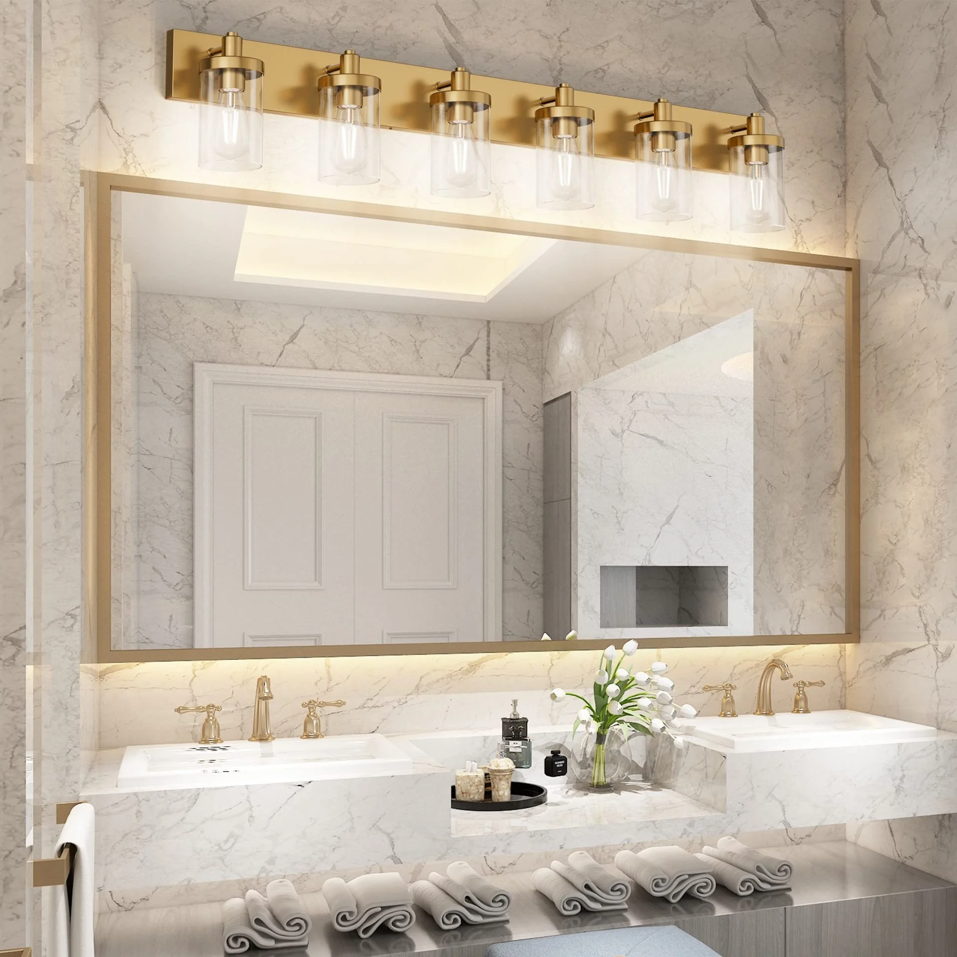A luxurious bathroom with a large mirror, marble walls, and a white countertop with two sinks. Above the mirror is a gold light fixture with multiple bulbs. On the countertop, there are bottles and a vase with white flowers. Below the countertop, rolled white towels are neatly stacked.