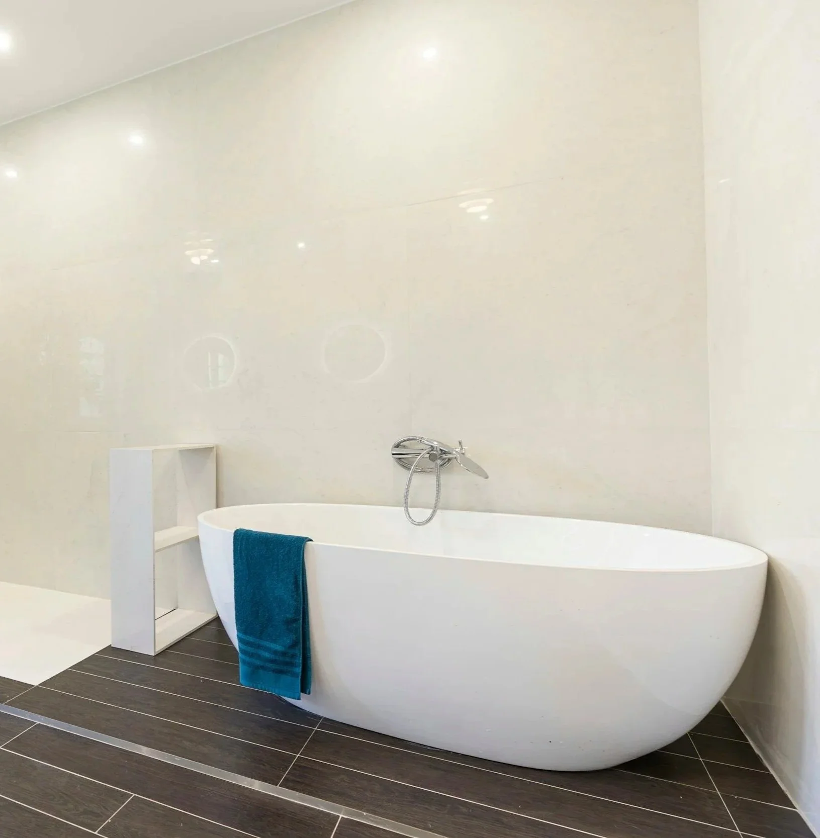 A modern bathroom featuring a white freestanding bathtub with a blue towel draped over the side. The wall behind the bathtub is smooth and white, with a handheld showerhead mounted above. The floor is tiled with dark, rectangular tiles with light grout lines, and a small white shelf stands next to the tub.