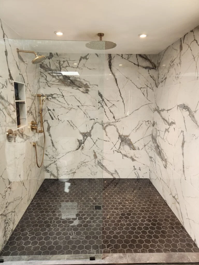 Modern walk-in shower with white marble walls featuring dark gray veining, a rainfall showerhead, a handheld shower, and a built-in wall niche, with dark hexagonal tile flooring.