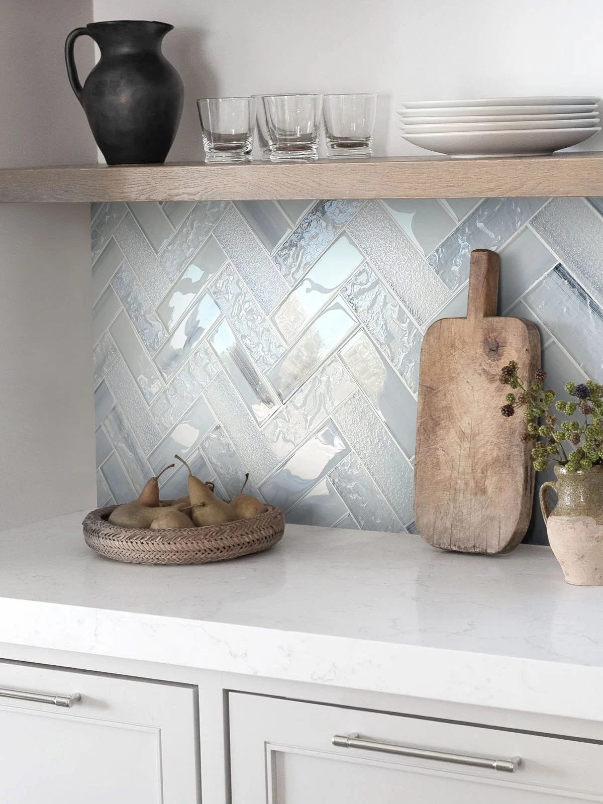 A kitchen countertop with a basket of pears, a wooden cutting board, a vase with flowers, and a tiled backsplash.