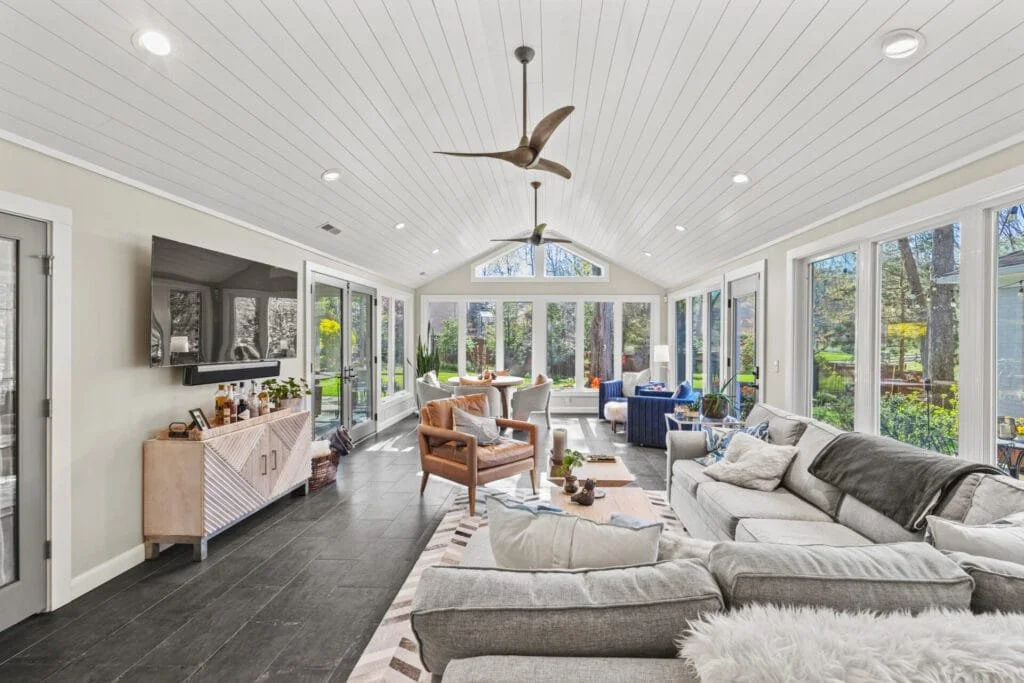 Bright sunroom with white shiplap ceiling, ceiling fans, large windows, and modern furniture.