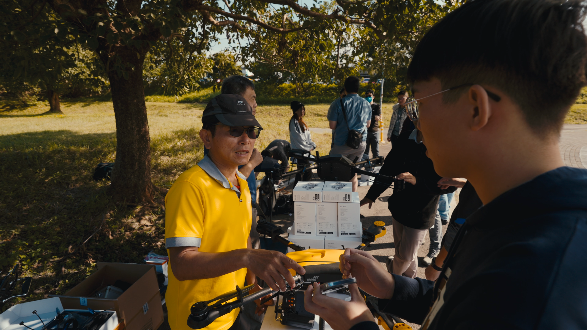 A man in a yellow shirt and black cap showing a drone controller to a young man with glasses at an outdoor event, with other people and trees in the background.