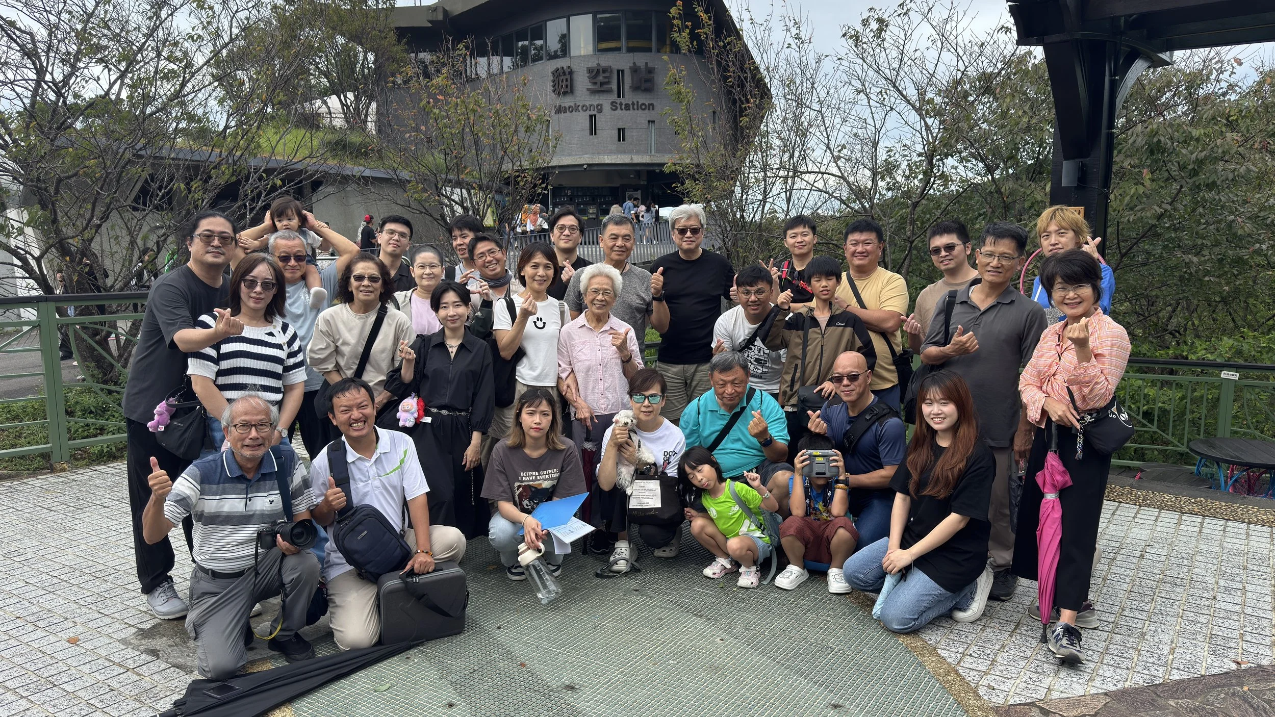 A large group of people posing for a photo in front of Maokong Station, with some making thumbs-up gestures. The group includes men, women, children, and an elderly woman, some holding cameras or bags, with a few children in front and adults behind. The station building with signage and trees is visible in the background.