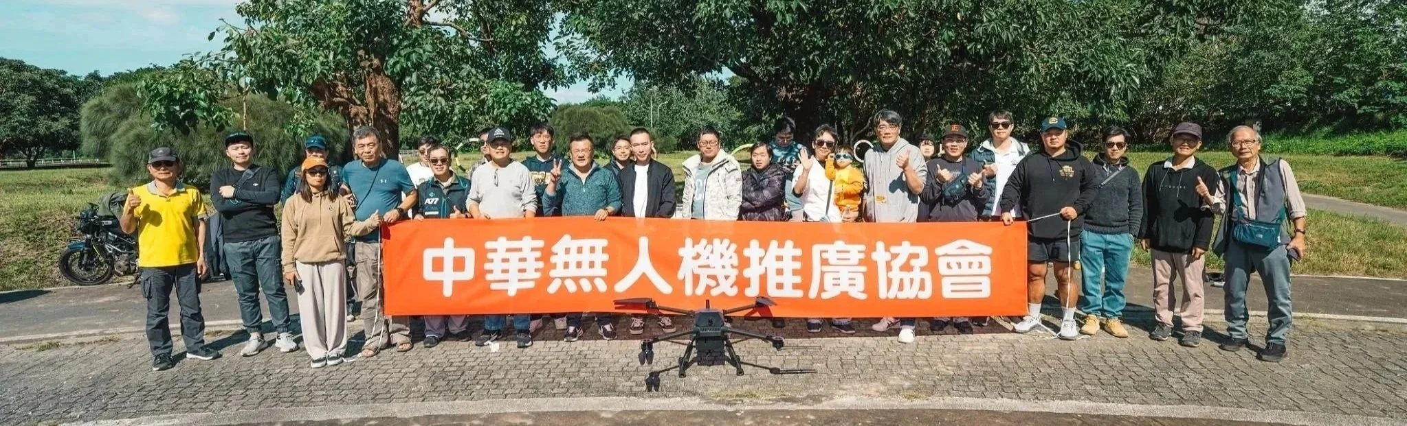 Group of people holding an orange banner with Chinese characters, outdoors on a sunny day, with trees and grass in the background.