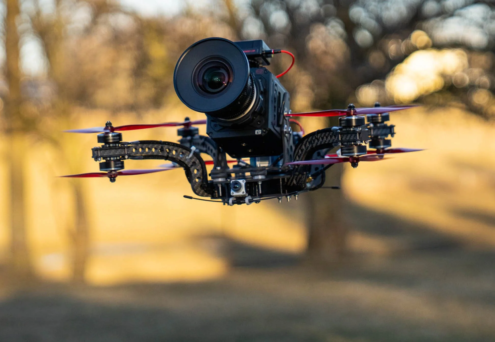 A drone with a mounted camera flying outdoors during golden hour with trees and sunlight in the blurred background.