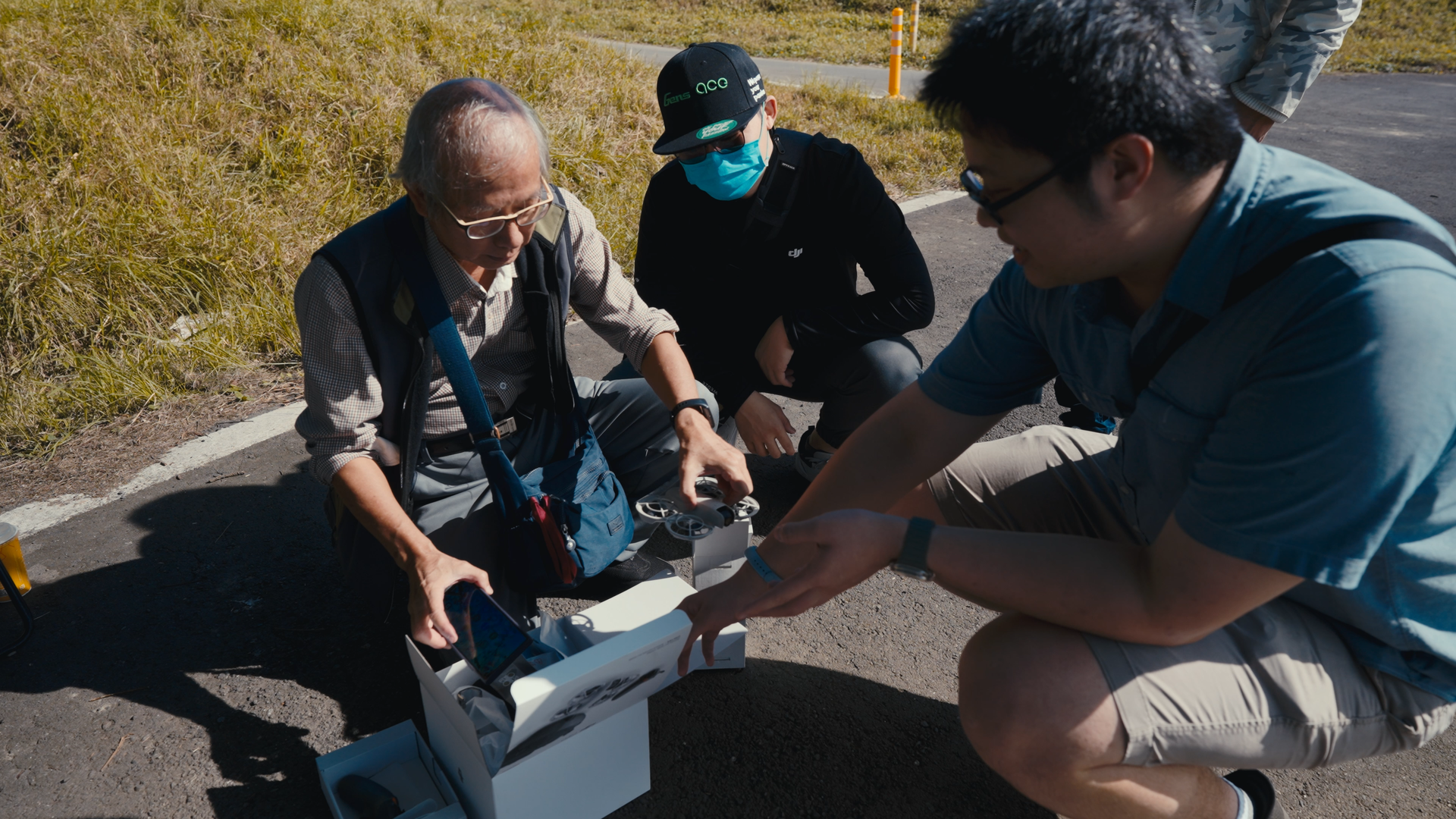 Three men examining a drone and a smartphone on a paved outdoor path next to a grassy area.