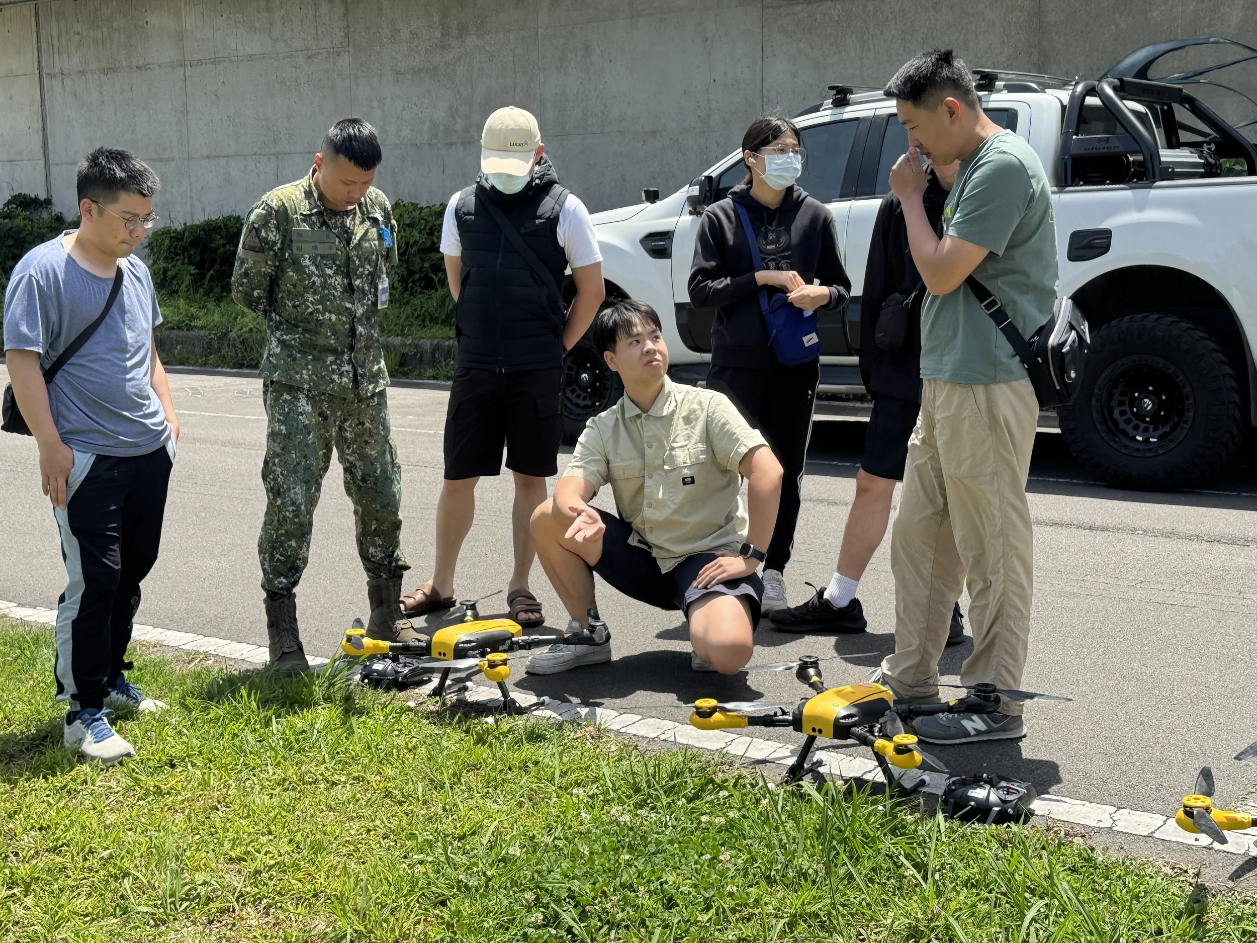Group of people, including military personnel, standing around a seated person, examining drone equipment on the ground during daytime.