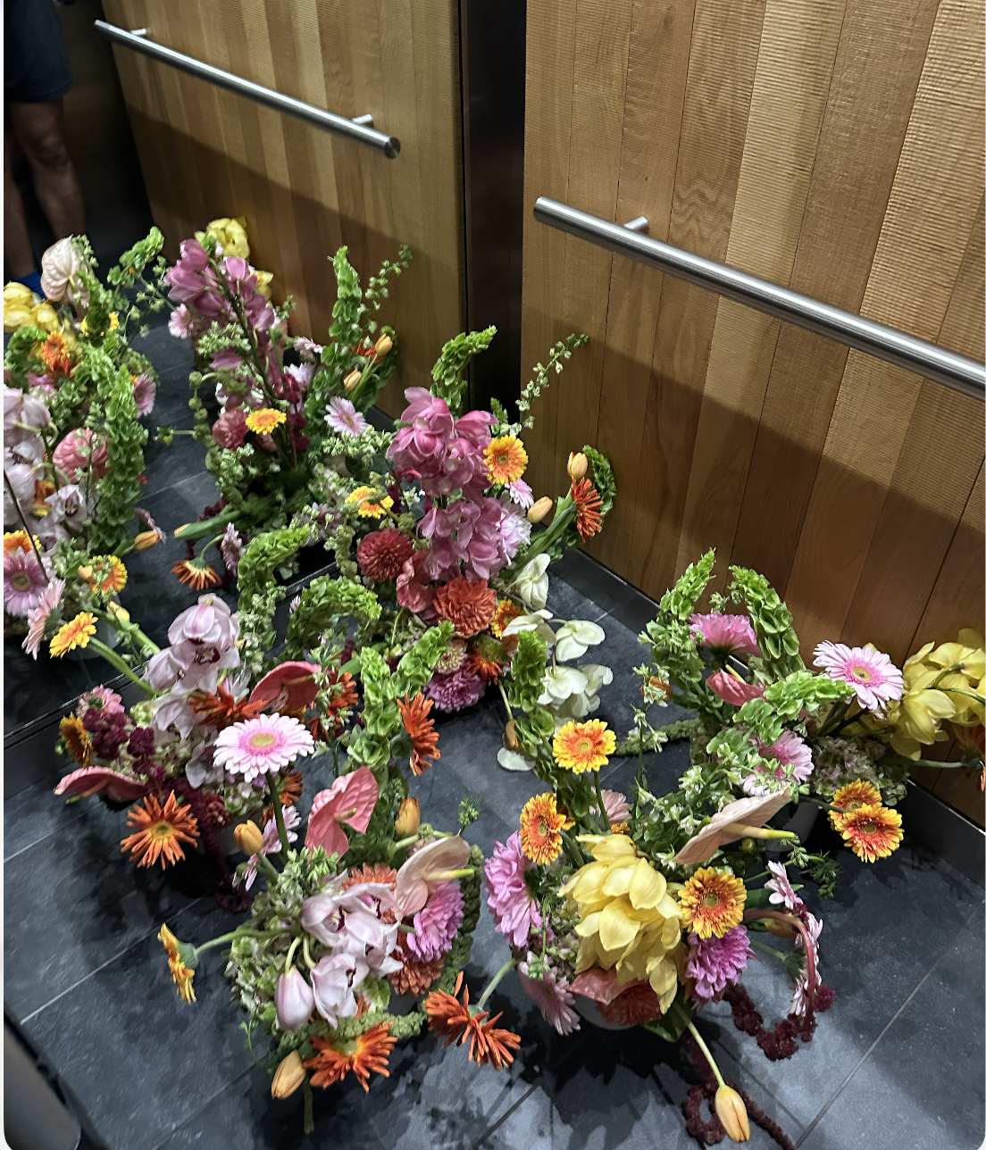 A collection of colorful flower arrangements with pink, yellow, orange, and white flowers placed on a dark tiled floor in front of a wooden wall inside an elevator.