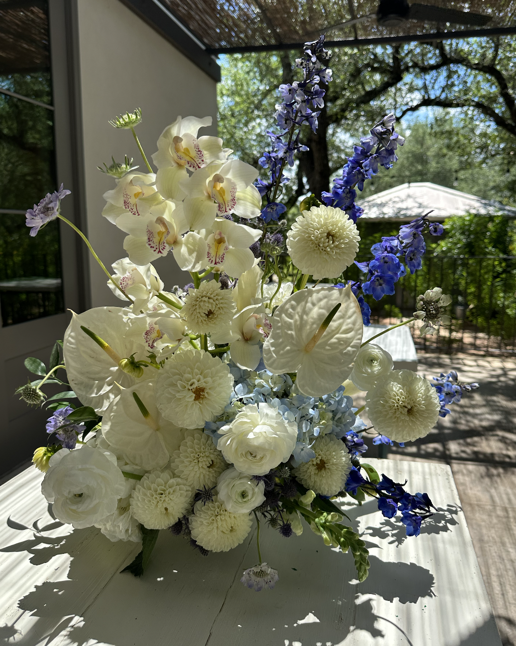 A bright flower arrangement with white, blue, and purple flowers on a white table, outdoors on a sunny day with trees and a deck in the background.