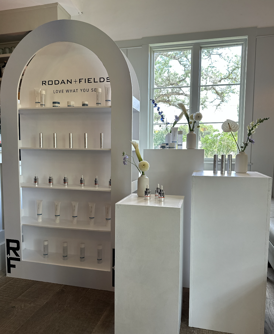 Display shelf with skincare products and vases with flowers near a window in a beauty or skincare store.