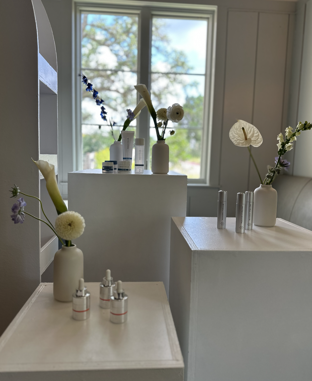 Inside a room with a large window, white shelves and tables display white vases with fresh flowers, skincare bottles, and small cosmetics containers, with a view of trees outside.