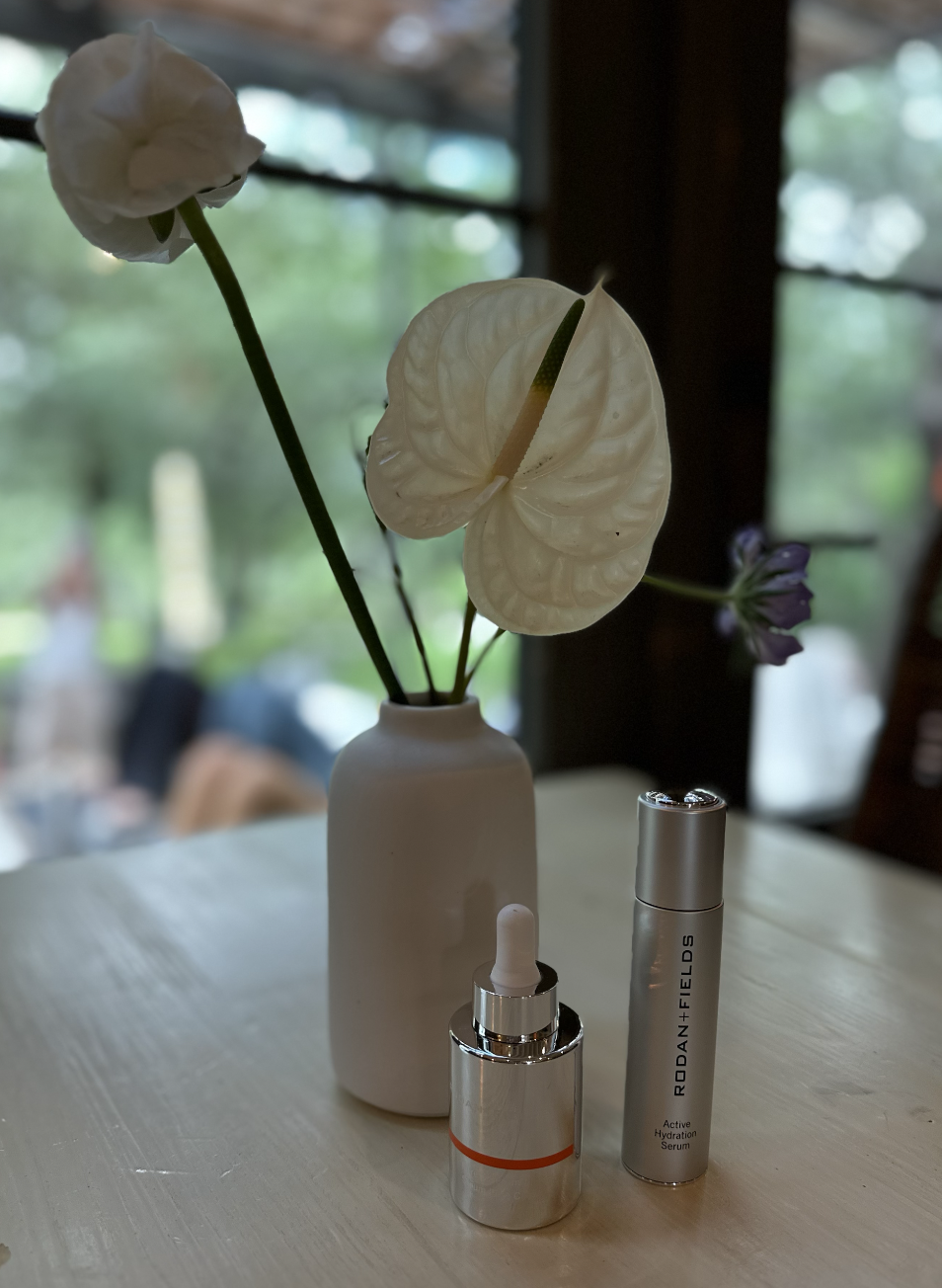 A white ceramic vase with anthurium flowers and a purple flower on a wooden table, with skincare serum bottles in front of it.