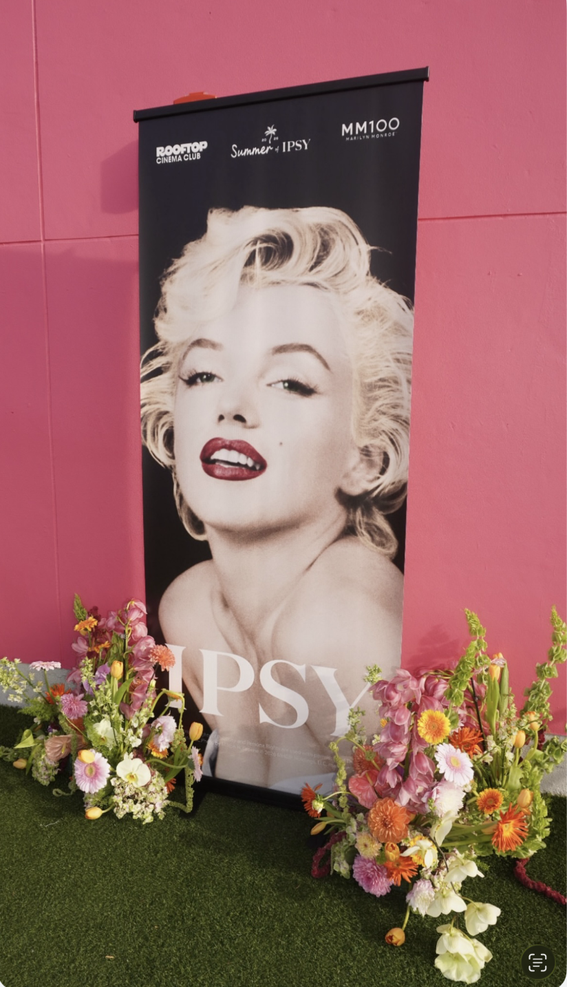 Poster of Marilyn Monroe with the word 'PSY' at the bottom, surrounded by flower arrangements, pink wall background.