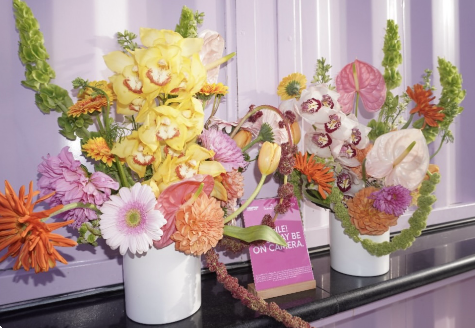 Colorful flower arrangements in white vases on a table with a purple background, including pink, orange, yellow, and white flowers.