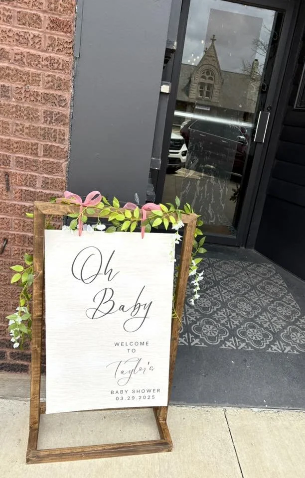 A white welcome sign with black cursive writing that says "Oh Baby" and "Welcome to Taylor's Baby Shower 03.29.2025," decorated with green leaves and pink ribbon, placed on a wooden easel outside a building with a brick wall and a glass door.