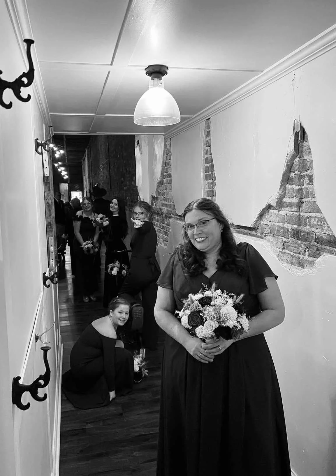 A woman holding a bouquet of flowers in a dress, smiling for the camera, with four other women in black dresses holding bouquets in the background, in a room with partially exposed brick walls.