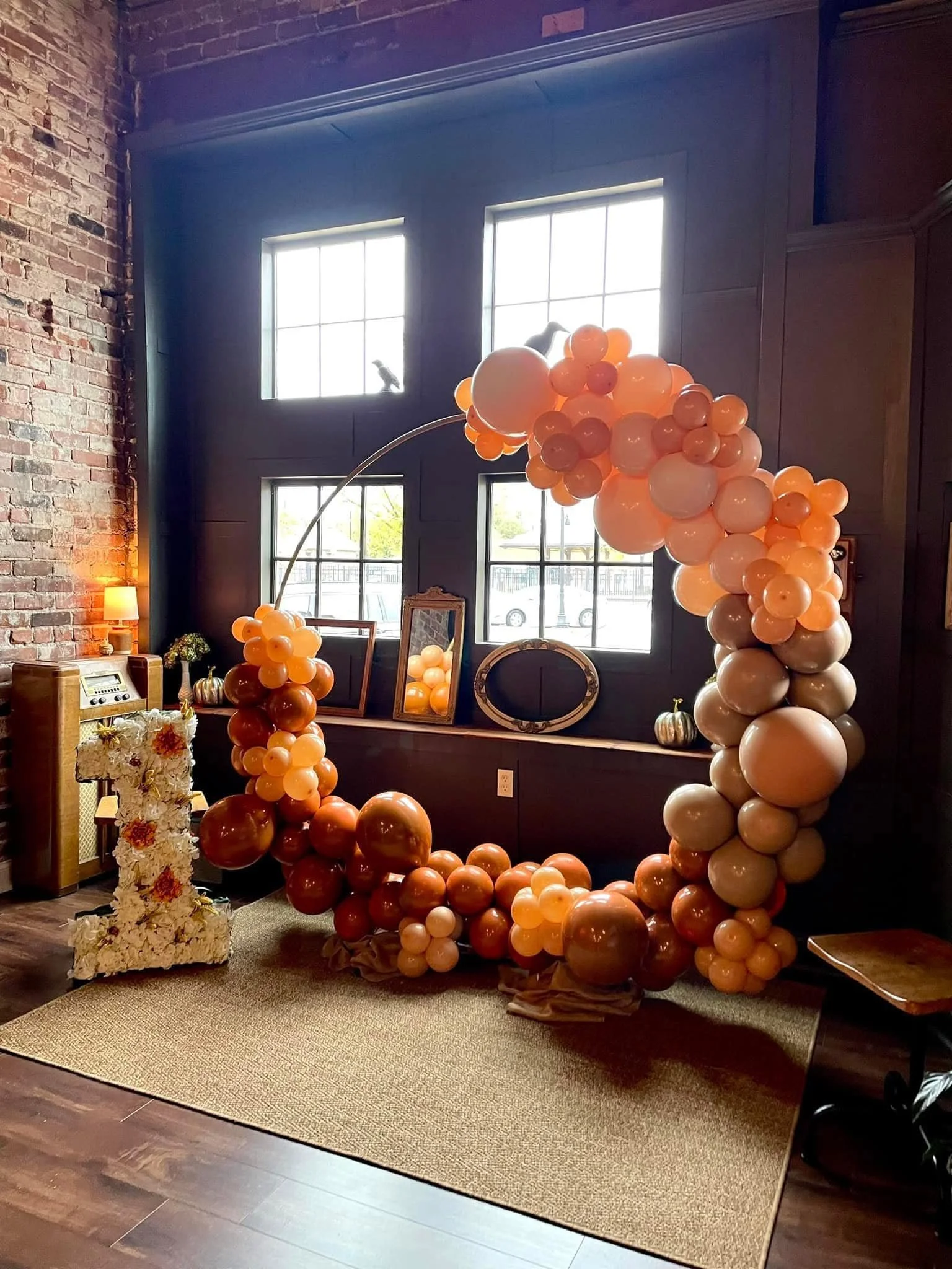 Indoor decoration featuring a large balloon arrangement in beige, peach, and brown tones forming a circular display with a rainbow-like shape. A white letter 'L' decorated with flowers is positioned on a beige rug in front of the balloons. The backgr