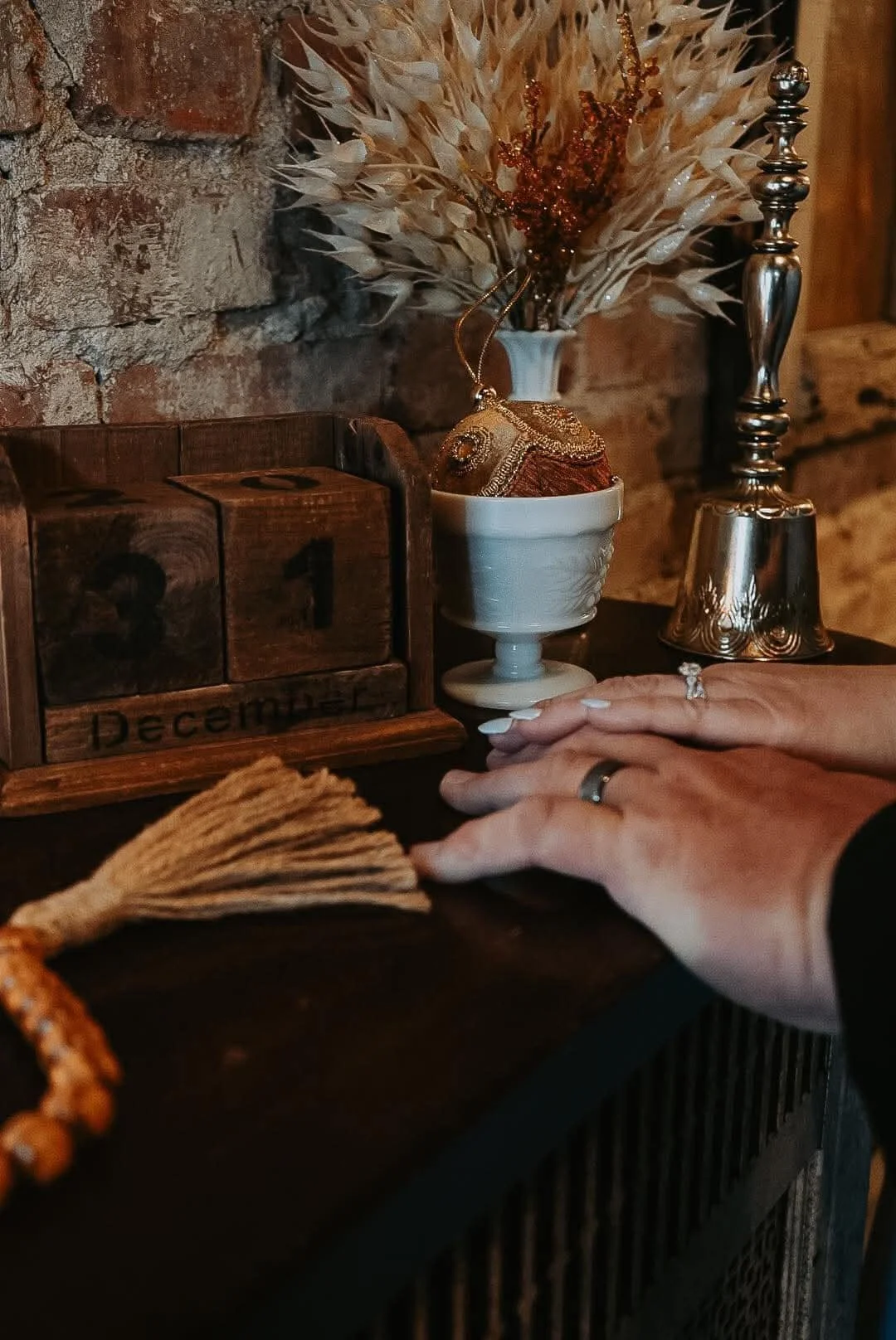 A person with rings on their fingers resting their hands on a dark wooden surface with decorative objects including a vintage calendar, a tassel, and a vase with dried flowers, against a brick wall background.